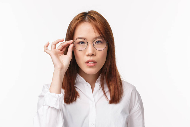 Κοντινό πλάνο πορτρέτο της ασιάτισσας όμορφης κοπέλας με λευκό πουκάμισο, δοκιμάζοντας νέα συνταγογραφημένα γυαλιά, διαλέγοντας γυαλιά στο κατάστημα οπτικών, κοιτάζοντας κάμερα αποφασισμένη, ακούγοντας σοβαρά να στέκεται λευκή - Φωτογραφία, εικόνα