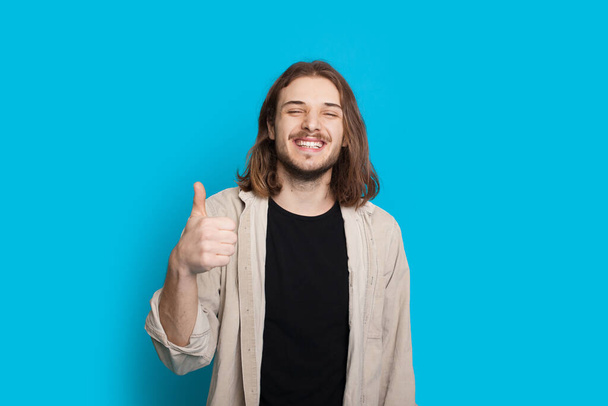 Bonito homem caucasiano com cabelos longos e barba está gesticulando o sinal semelhante em um fundo azul
 - Foto, Imagem