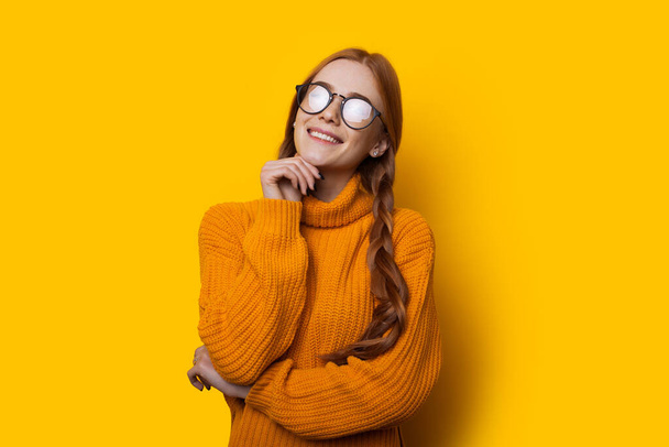 Jengibre dama caucásica con pecas con gafas está tocando su barbilla sobre un fondo amarillo
 - Foto, Imagen