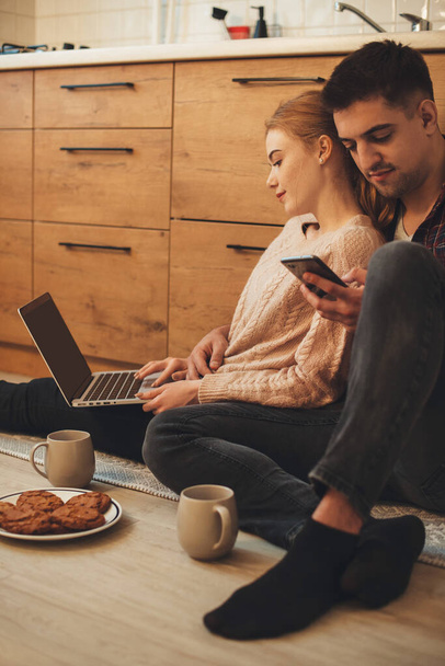 Adorável casal caucasiano sentado na cozinha no chão navegando em um laptop e telefone enquanto toma um café da manhã
 - Foto, Imagem