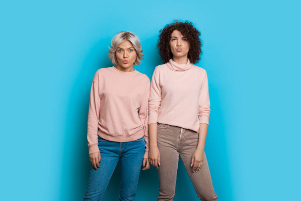 Two caucasian sisters with curly hair wearing the same sweater are posing and gesturing with lips a kiss on a blue background - Photo, Image