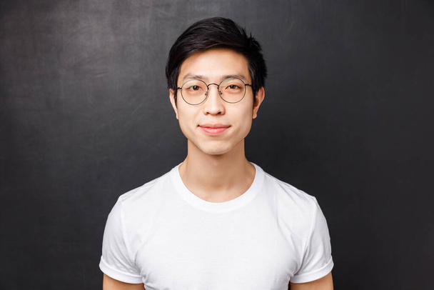 Close-up portrait of simple cute asian man in white t-shirt and glasses, smiling at camera looking friendly and relaxed, posing over black background alone, lifestyle and people concept - Foto, Imagem