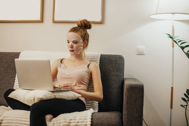 Caucasian woman with red hair is wearing a cleaning mask on her face while sitting on the sofa and using a laptop - Fotografie, Obrázek