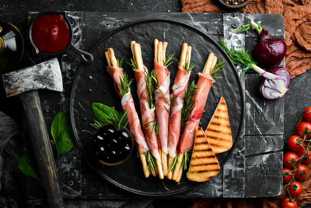 Italian antipasto. Bread sticks grissini with prosciutto ham on a black plate. Rustic style. Top view. Free space for your text. - Foto, afbeelding