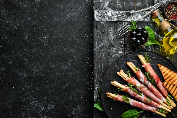 Italian antipasto. Bread sticks grissini with prosciutto ham on a black plate. Rustic style. Top view. Free space for your text. - Fotografie, Obrázek