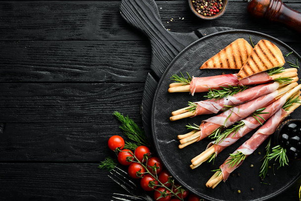 Italian antipasto. Bread sticks grissini with prosciutto ham on a black plate. Rustic style. Top view. Free space for your text. - Photo, Image