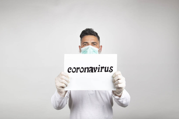 Cerrahi maskeli bir adamın portresi beyaz arka planda "koronavirüs" yazan beyaz bir tabela tutuyor.. - Fotoğraf, Görsel