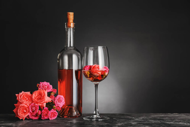 Pullo viiniä, lasia ja ruusuja pöydällä tummaa taustaa vasten
 - Valokuva, kuva