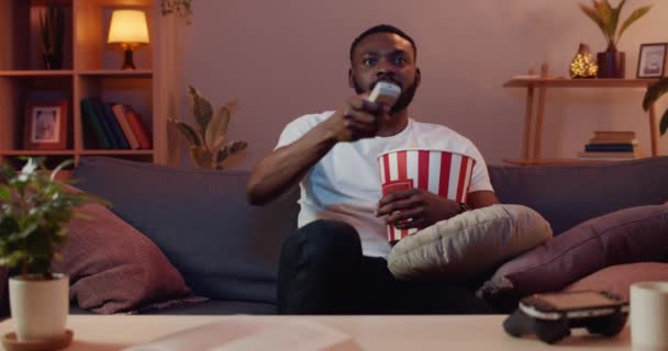 Handsome guy using remote control while sitting on sofa with popcorn bowl and eating. Relaxed young man watching film on TV and enjoing free time at home. Concept of leisure. - Πλάνα, βίντεο
