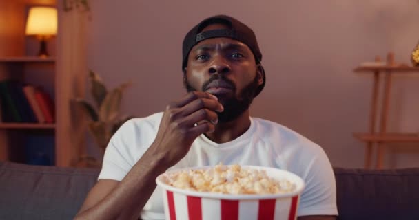 Handsome young guy making different face expressions while watching interesting film at home. Excited african eating popcorn from paper bowl while sitting on sofa in front of TV. - Séquence, vidéo