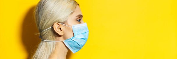 Retrato del perfil del estudio de una joven rubia con ojos azules, con máscara de gripe médica, pequeña protección contra el coronavirus. Aislado sobre fondo amarillo con espacio de copia
. - Foto, Imagen