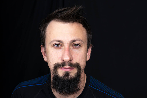 een close-up portret van een jonge man met baard in donkere kleren op een donkere achtergrond - Foto, afbeelding