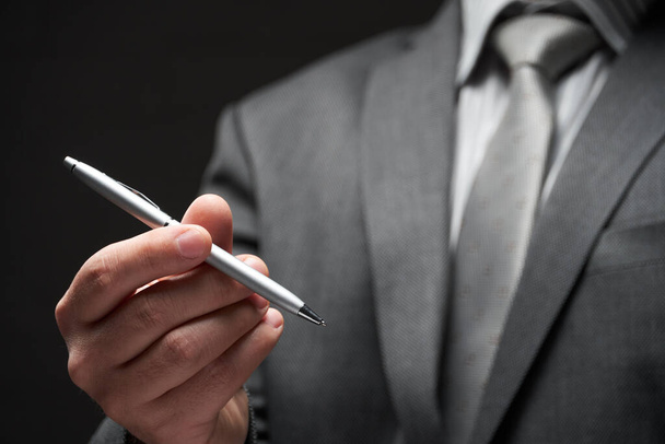 Stift-Nahaufnahme in der Hand eines Geschäftsmannes, grauer Anzug, dunkler Wandhintergrund - Foto, Bild