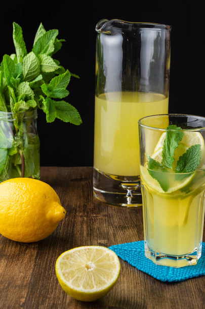 Вид зі скла і глечика з лимонним соком на темному дерев'яному столі з лимоном, половиною листя лимона і м'яти з чорним тлом вертикально
 - Фото, зображення