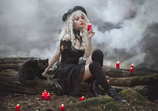Witch girl in a hat with a black cat in smoke holds red candles. Sexy witch in the forest. - Foto, Imagen