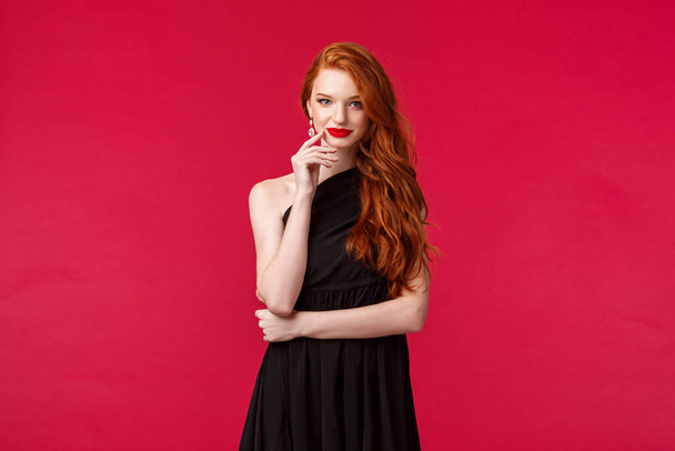 Elegance, fashion and woman concept. Portrait of seducative good-looking redhead woman in slim elegant black dress, red lipstick and evening makeup, look daring and sassy, red background - Photo, Image