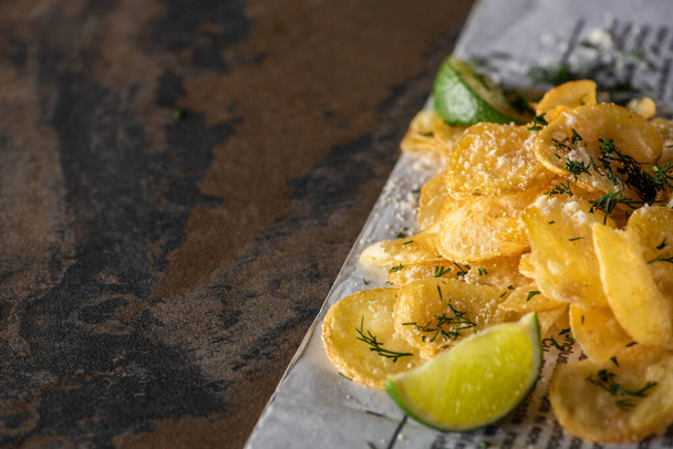 selective focus of salty potato chips near sliced lime and newspaper on marble surface  - Photo, Image