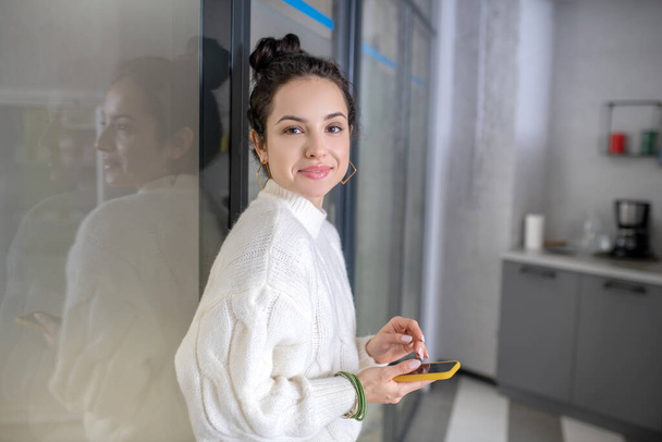 Young woman leaning on glass wall, holding mobile, smiling - Valokuva, kuva