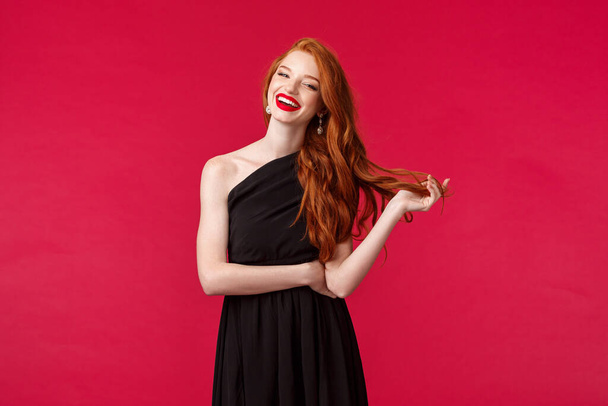 Elegance, fashion and woman concept. Portrait of confident sexy and stylish young redhead woman in red lipstick, evening makeup, wearing elegant black dress, laughing at formal event, red background - Photo, Image