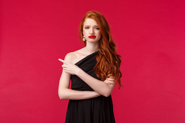 Portrait of skeptical, picky elegant redhead girlfriend in stylish black dress, grimacing disappointed, pointing finger left at something bad, dislike and disagree, stand red background - Photo, Image