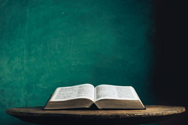 Open Holy Bible on a old round wooden table. Beautiful green wall background. - Photo, Image