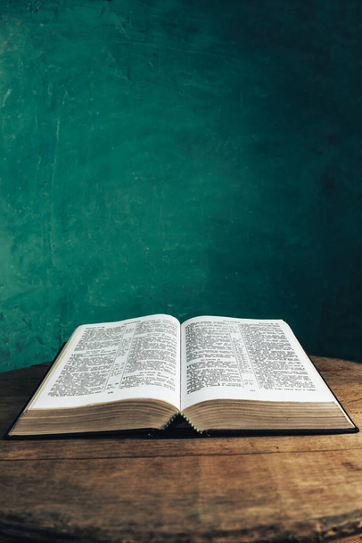 Open Holy Bible on a old round wooden table. Beautiful green wall background. - Photo, Image