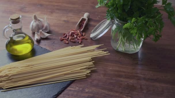 典型的なイタリア料理のスパゲッティ・アリオ・オリオ・エ・ペペロンチーノ(ニンニク、オイル、唐辛子)材料の上から下まで、木製のテーブルの上にパセリの小枝とガラス瓶 - 映像、動画