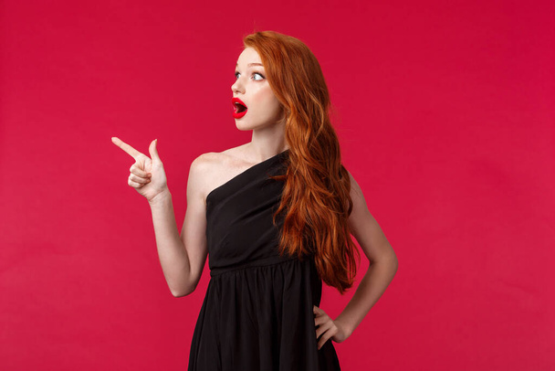 Verrast en verbaasd jonge roodharige vrouw in elegante partij zwarte jurk, vallen kaak pinnen als staren en wijzende vinger links naar iets schokkende, staande rode achtergrond - Foto, afbeelding