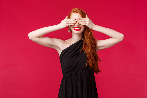 Fashion, luxury and beauty concept. Portrait of elegant gorgeous young woman with red long hair, black luxurious dress, cover eyes blindfolded counting ten to see surprise, smiling, red background - Photo, Image