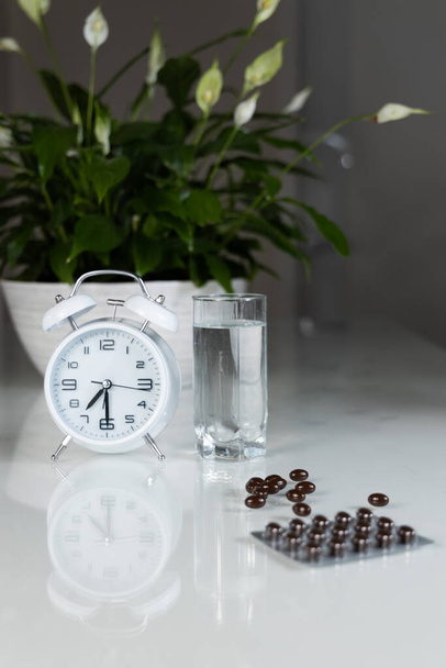 White alarm clock and medical pills with a glass of water on the table. Healthcare and medicine. Vitamins and mineral supplements in capsules. Vertical shot. - Fotoğraf, Görsel