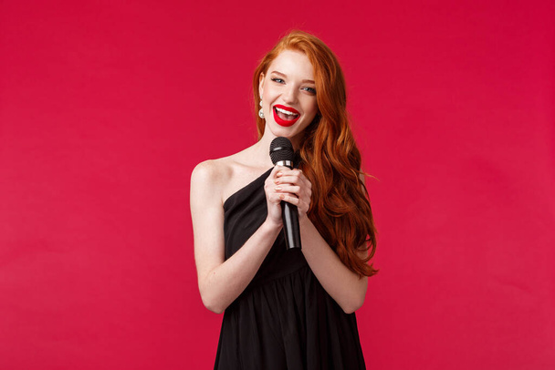 Retrato de una hermosa mujer joven femenina vestida de negro, sosteniendo el micrófono cantando karaoke o interpretando canciones frente a la audiencia en un evento público, de pie fondo rojo
 - Foto, imagen