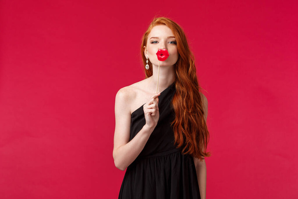 Portrait of feminine good-looking redhead woman in black stylish dress, holding fake lips cardboard over mouth as if give passionate kiss, looking at camera tender, stand red background - Photo, Image