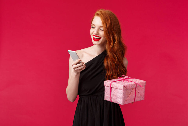 Portrait de joyeuse jeune femme rousse élégante en robe élégante noire, tenant boîte cadeau et smartphone, SMS ami merci comme recevoir présent par courrier, stand fond rouge
 - Photo, image