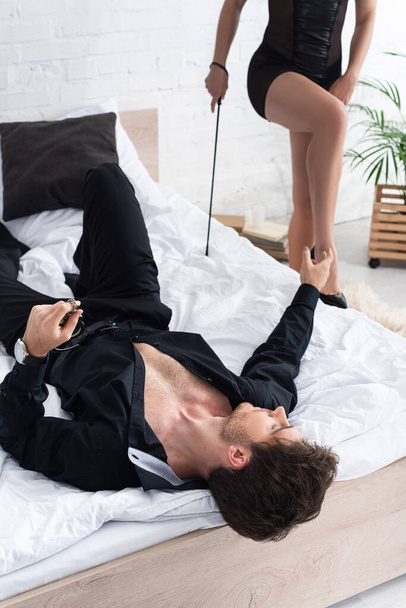 Επιλεκτική εστίαση της γυναίκας με μαστίγωμα μαστίγιο και ο άνθρωπος που βρίσκεται στο κρεβάτι με απλωμένο το χέρι στην κρεβατοκάμαρα - Φωτογραφία, εικόνα