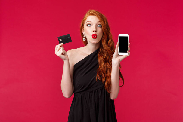 Γοητευμένος και ενθουσιασμένος όμορφος ευρωπαϊκή γυναίκα με μακριά σγουρά μαλλιά τζίντζερ, φορούν μαύρο φόρεμα, κρατώντας το κινητό τηλέφωνο, δείχνει οθόνη smartphone και πιστωτική κάρτα, ψώνια και internet έννοια - Φωτογραφία, εικόνα