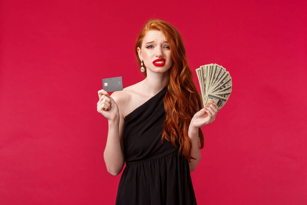 贅沢、美しさとお金の概念。多くの現金ドルとクレジットカードを保持している決定的ではなく、よく見えない若い赤毛のガールフレンドは、不確定な悲しみはそれがどのように無駄に疑問を持っています - 写真・画像