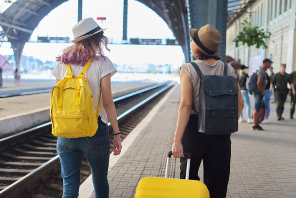 Railway station, people passengers with luggage. Two traveling women with suitcase backpacks walking along platform, back view - Photo, image