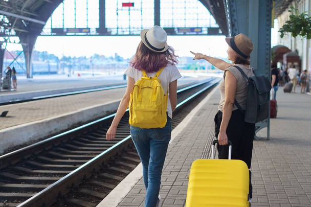 Railway station, people passengers with luggage. Two traveling women with suitcase backpacks walking along platform, back view - Фото, зображення