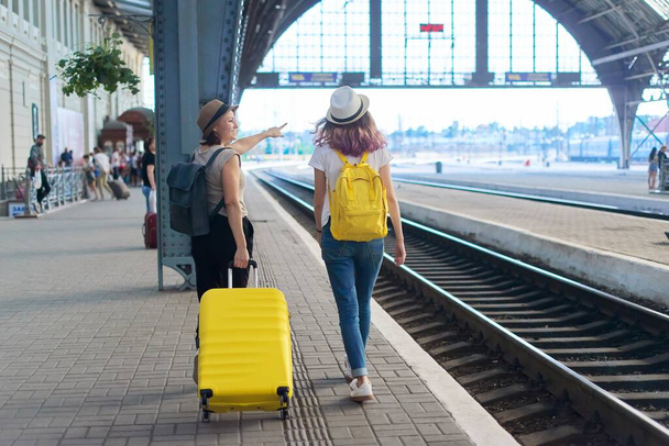 Railway station, people passengers with luggage. Two traveling women with suitcase backpacks walking along platform, back view - Zdjęcie, obraz