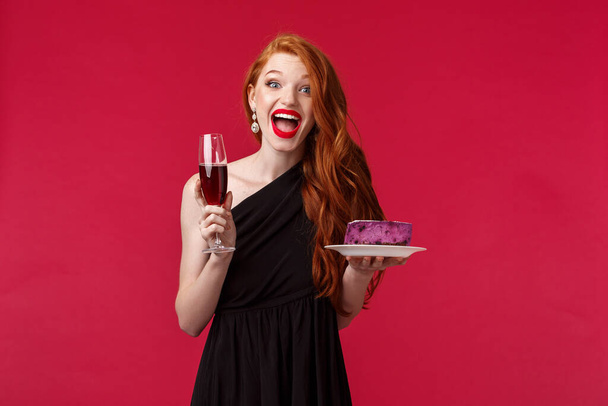 Портрет чудової святкової і схвильованої жінки, яка святкує день народження, відчуває себе задоволеною і щасливою, тримає скляне шампанське і б-денний торт, розважається, одягається чорна елегантна сукня на червоному тлі
 - Фото, зображення