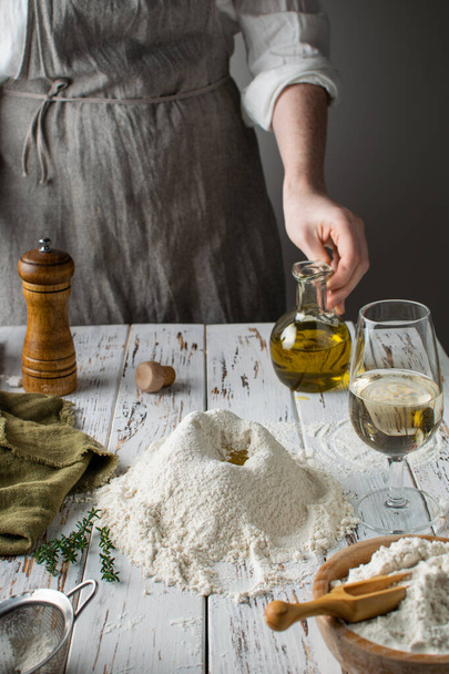Process of preparation homemade pasta or bread, female hande holding glass bottle with olive oil into flour, rustic white wooden table - Foto, imagen