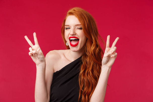 Close-up portrait of sassy and carefree redhead girl having fun, enjoying awesome party, show kawaii peace signs dancing joyfully, celebrating holiday in black dress and red lipstick - Photo, image