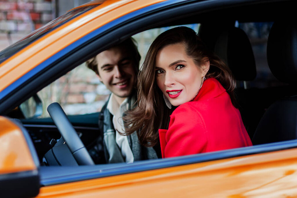 Mooi meisje portret in een rode jas poseren in oranje auto met een jonge man die uit focus. meisje in een jas buiten. mooi meisje in een modieuze casual jas. lenteherfstcollectie. - Foto, afbeelding