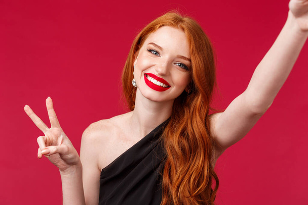 Close-up portrait of beautiful redhead european woman celebrating her birthday or enjoying party, taking selfie with peace sign and beaming satisfied smile, having fun, red background - Photo, Image