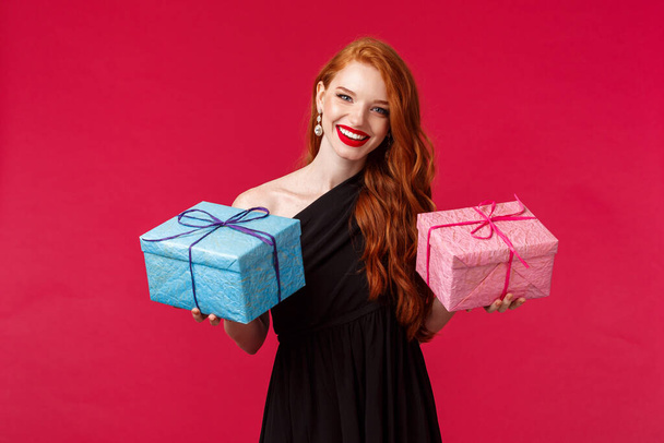 Celebration, holidays and women concept. Portrait of elegant gorgeous young redhead woman in black party dress, holding two gifts in blue and pink boxes, smiling camera cheerful, red background - Photo, Image