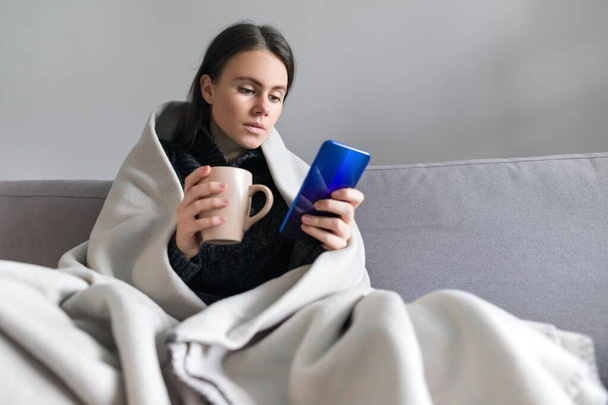 Young woman with cup, smartphone, sitting at home on sofa under warm fleece blanket, copy space - Φωτογραφία, εικόνα