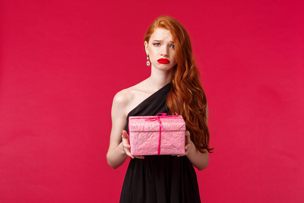 Celebration, holidays and women concept. Portrait of gloomy and upset birthday girl feel distressed and uneasy, holding one pink box with b-day gift, sobbing, no one came to party, red background - Photo, image