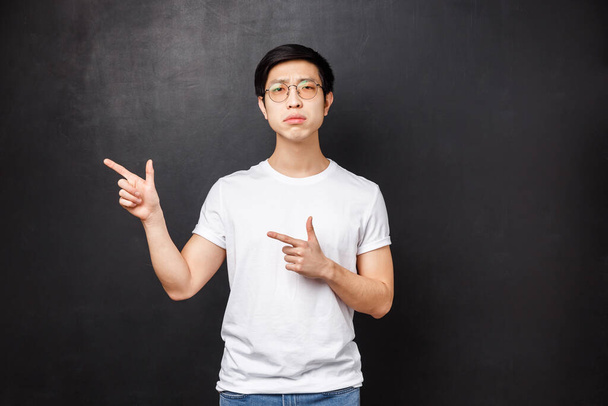 Προσωπογραφία δύσπιστου και αμφίβολου νεαρού σοβαρού έξυπνου άντρα που σκέφτεται πρόταση έχοντας αμφιβολίες, δείχνοντας τα δάχτυλα αριστερά στη διαφήμιση και κοιτώντας την κάμερα καχύποπτη με δυσπιστία, μαύρο φόντο - Φωτογραφία, εικόνα