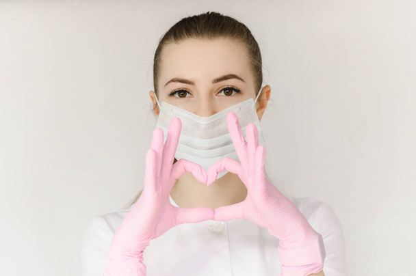 young successful girl doctor in protective mask and white coat shows sign with heart hand  - 写真・画像