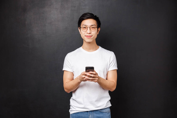 Technologie, Gadgets und Menschen-Konzept. Schöner junger entschlossener asiatischer Typ in T-Shirt und Brille mit Handy-App, Smartphone in der Hand und in die Kamera lächeln, schwarzer Hintergrund - Foto, Bild